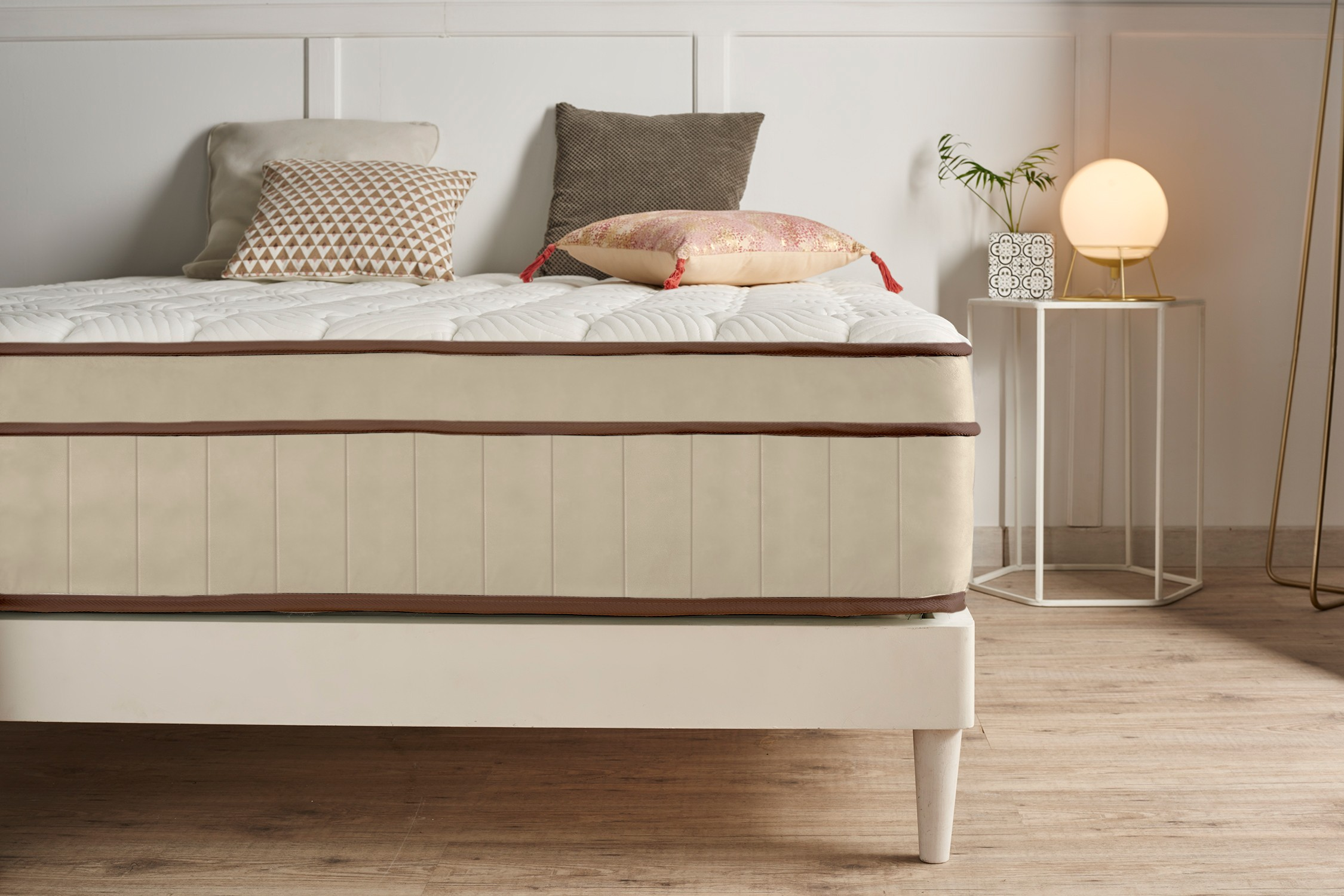 Un colchón grueso en una cama blanca con cojines de colores y una lámpara de noche al fondo.
