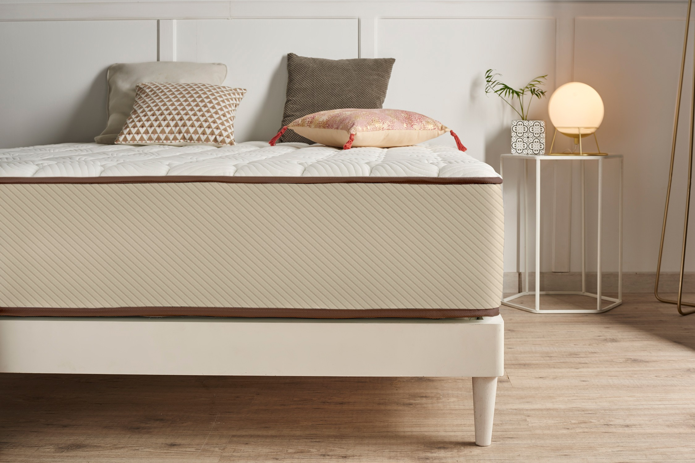 Una cama blanca con un colchón beige, cojines coloridos y una lámpara de noche.