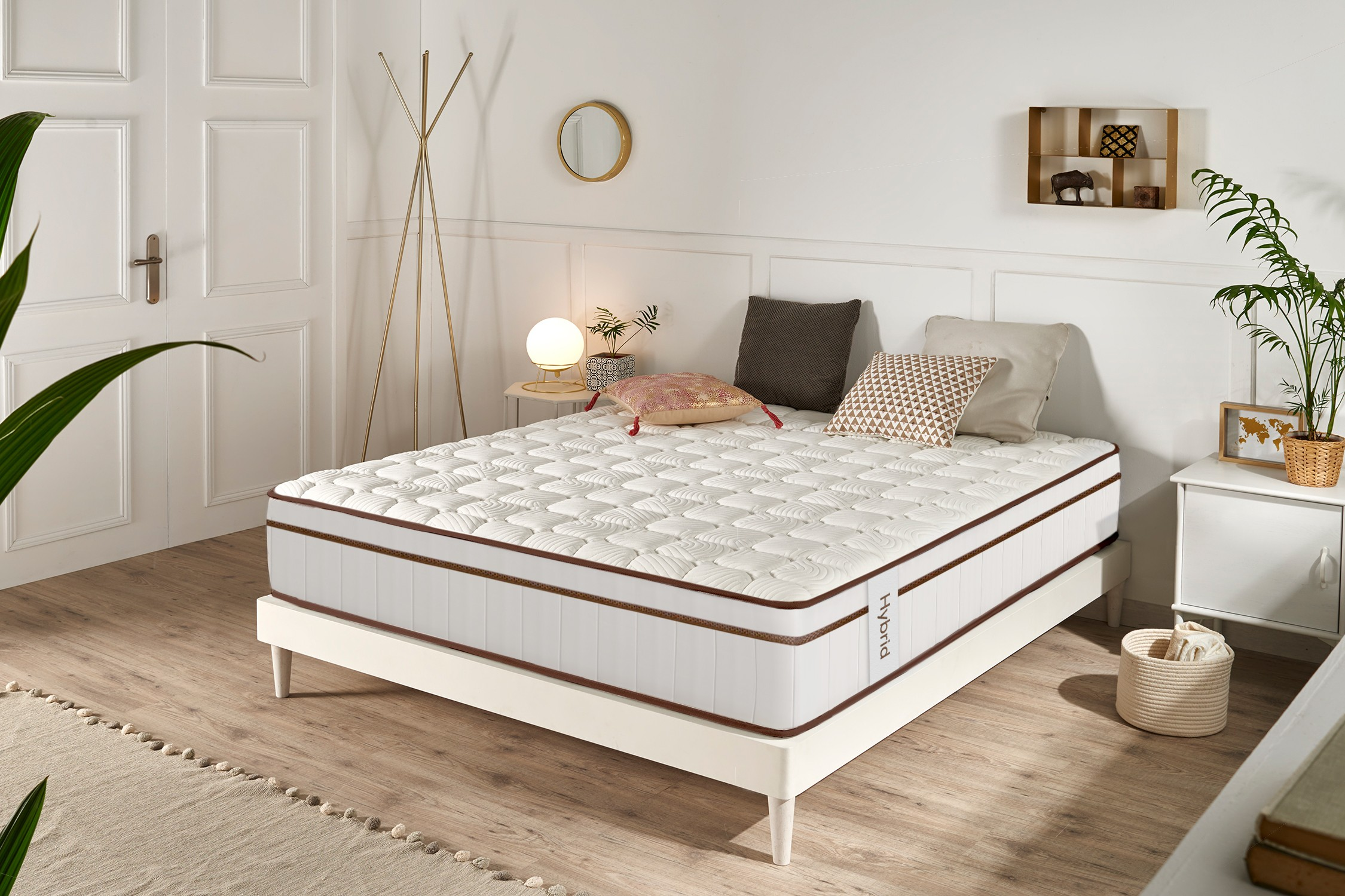 Un colchón híbrido blanco en una base de madera en un dormitorio luminoso con plantas, cojines y una mesita.