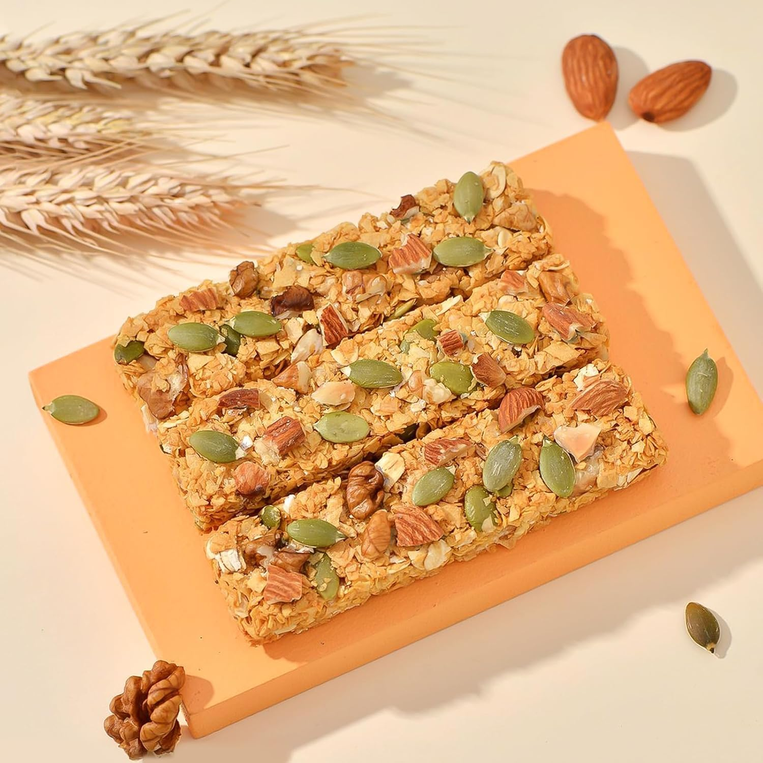 Drei Hafer-Nuss-Müsliriegel auf einem orangefarbenen Brett, belegt mit Kürbiskernen, Walnüssen und Mandeln, mit Weizenähren.