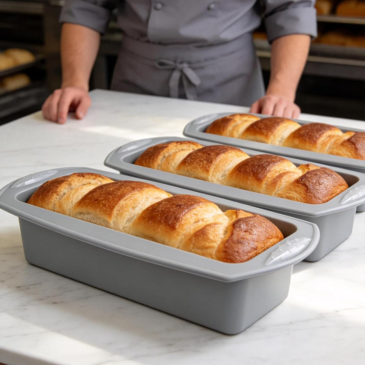 Ein Bäcker in grauer Uniform steht hinter drei grauen Kastenformen mit frisch gebackenem, goldbraunem Brot.