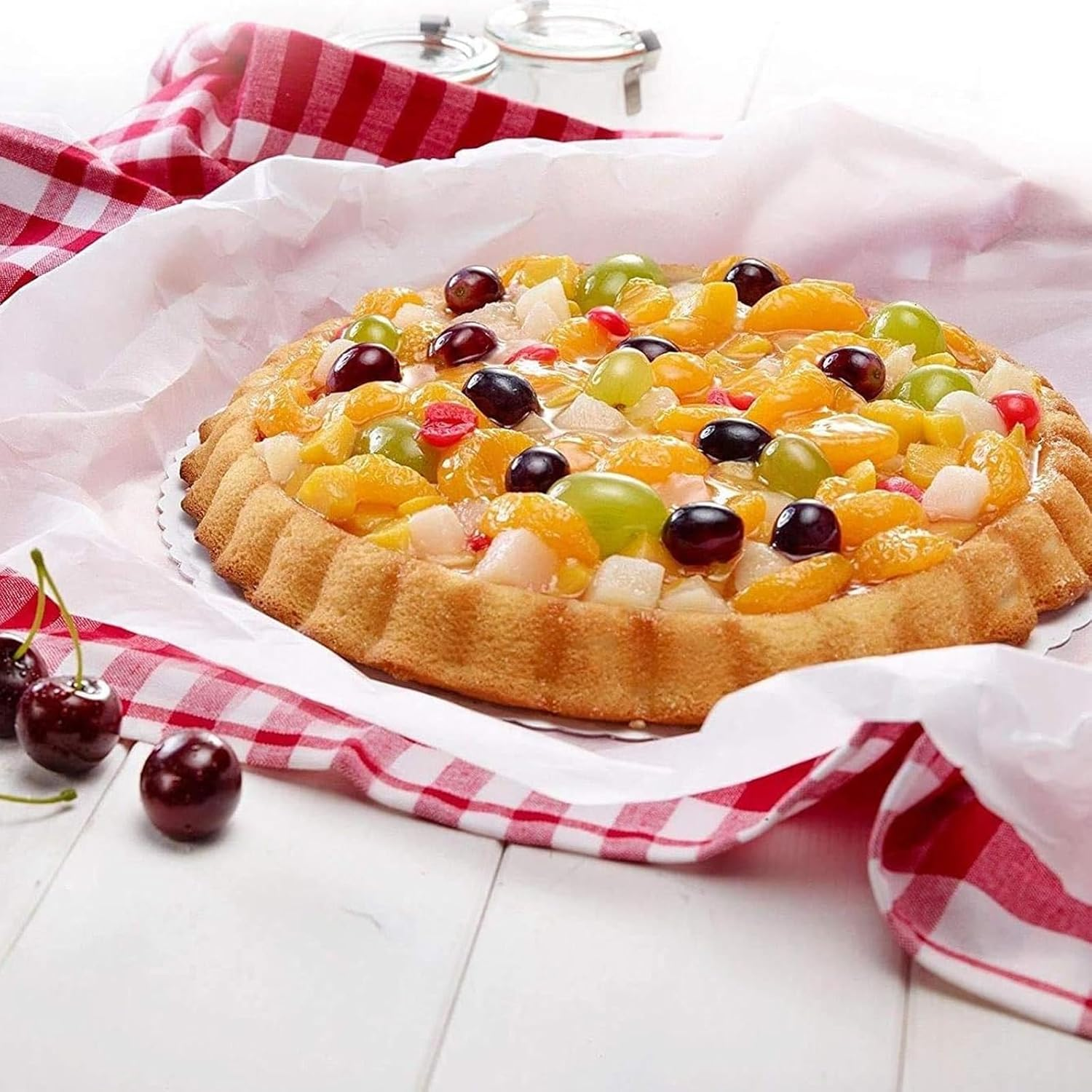 Ein frischer Obstkuchen mit Trauben, Mandarinen und Kirschen auf weißem Backpapier und rot-weiß kariertem Tuch.