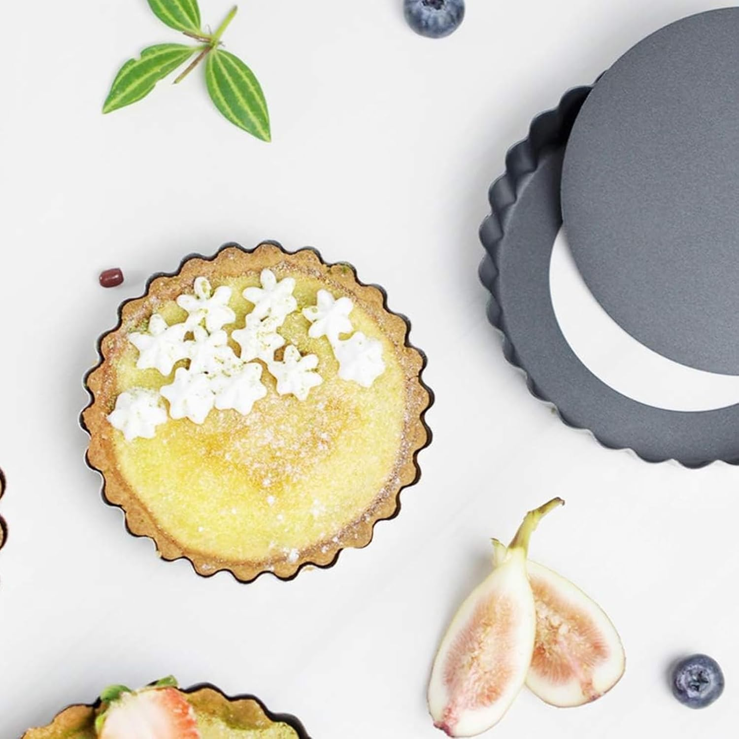 Ein gebackener Kuchen mit Sahneblumen, daneben eine Tarteform, Feigen und Blaubeeren auf weißem Grund.