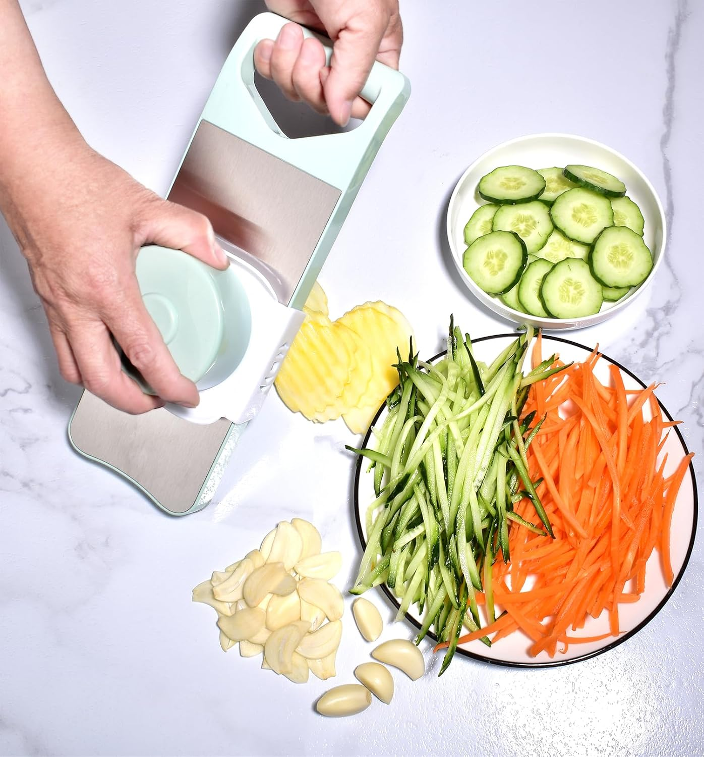 Hände schneiden Gemüse mit einem grünen Gemüsehobel auf weißer Oberfläche neben Schüsseln mit Gurken, Karotten und Knoblauch.