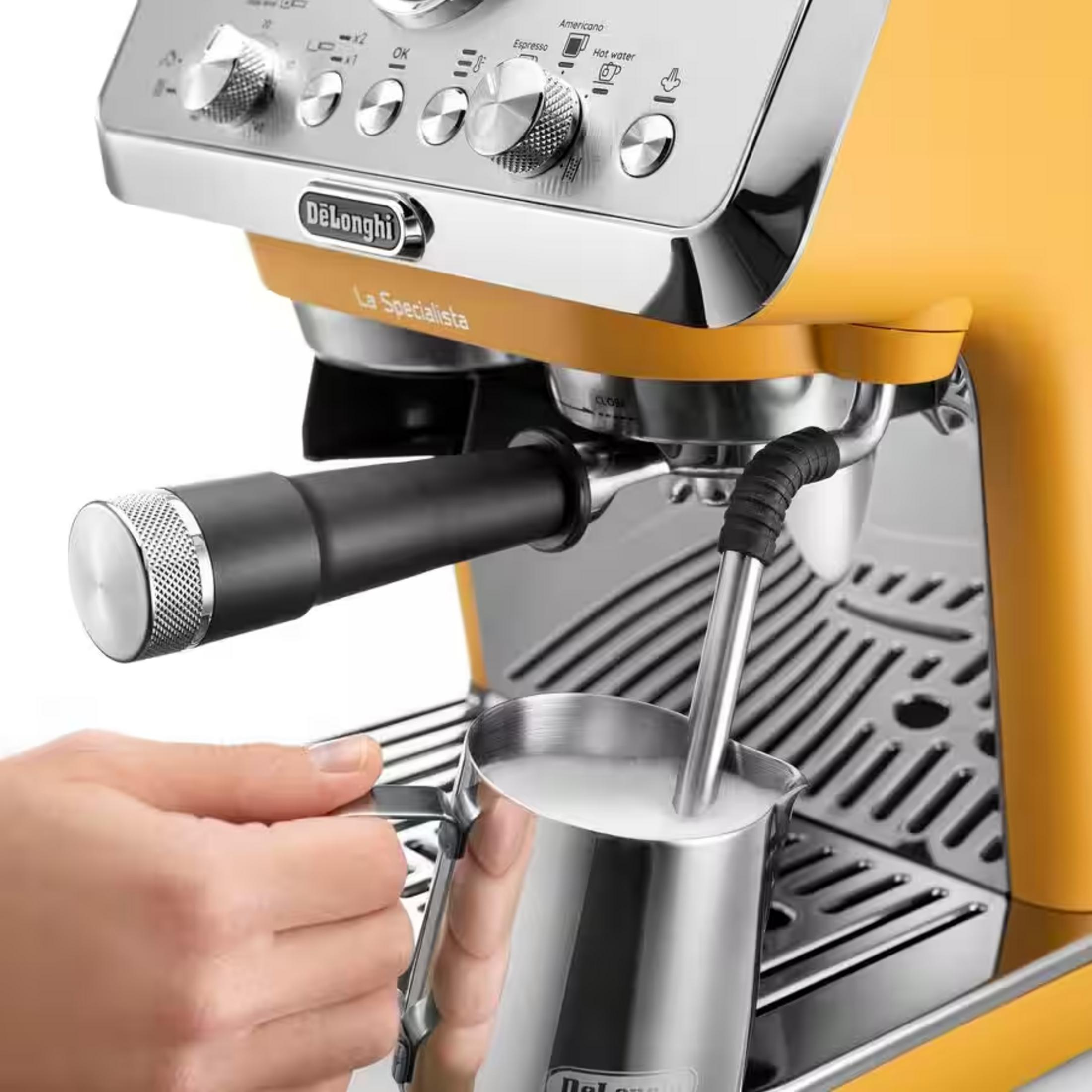 Bir el, sarı bir DeLonghi espresso makinesinin buhar çubuğunun altında bir süt sürahisi tutuyor.
