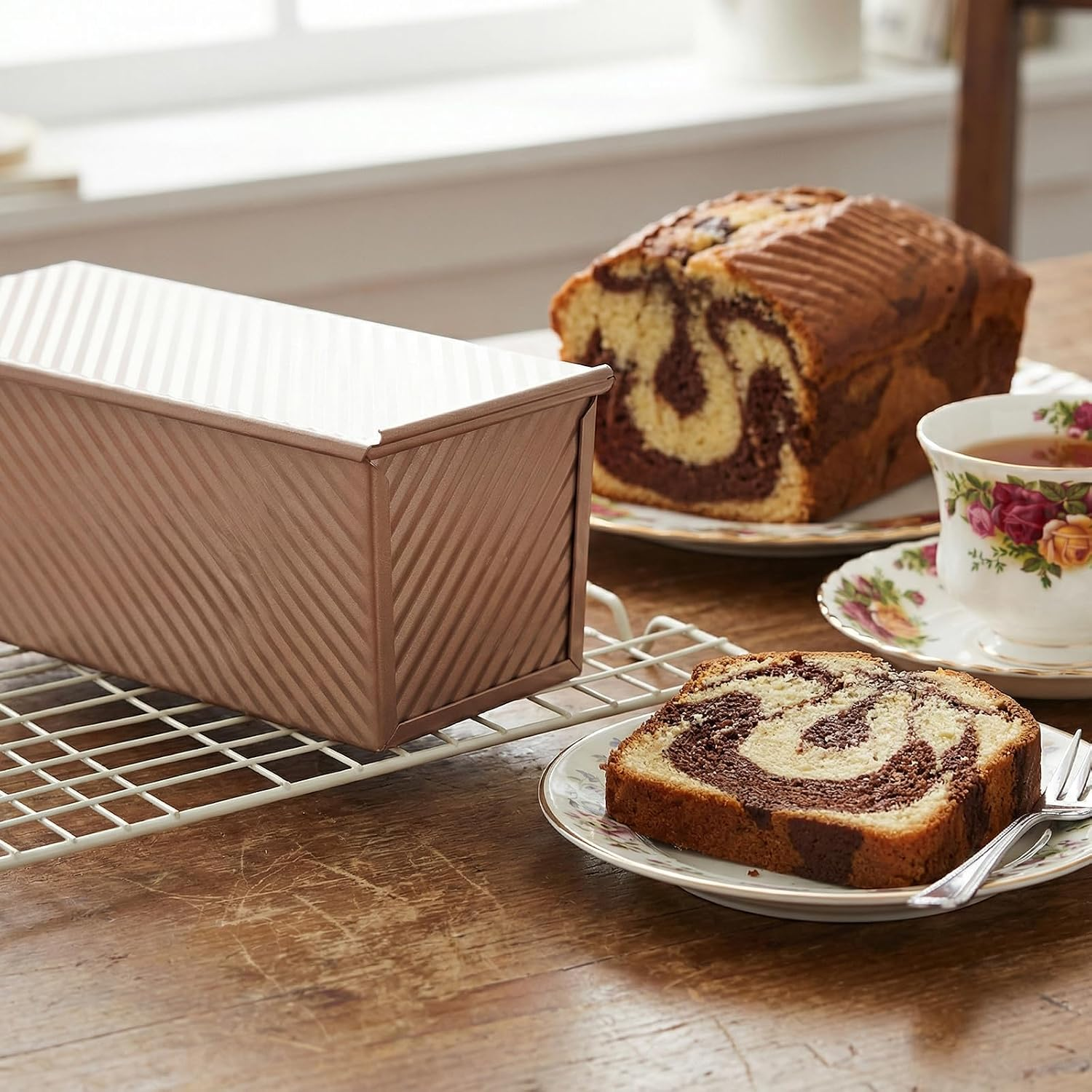 Eine rosa Kastenform steht auf einem Gitter, daneben Marmorkuchen auf einem Teller und eine Teetasse.