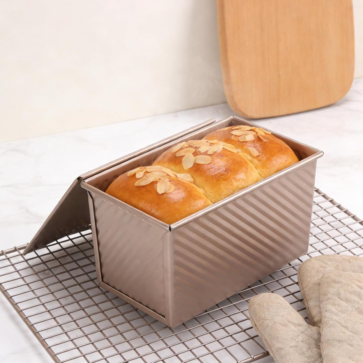 Ein längliches Brot mit Mandelblättchen in einer Metallbackform auf einem Kühlgitter, daneben ein Ofenhandschuh.