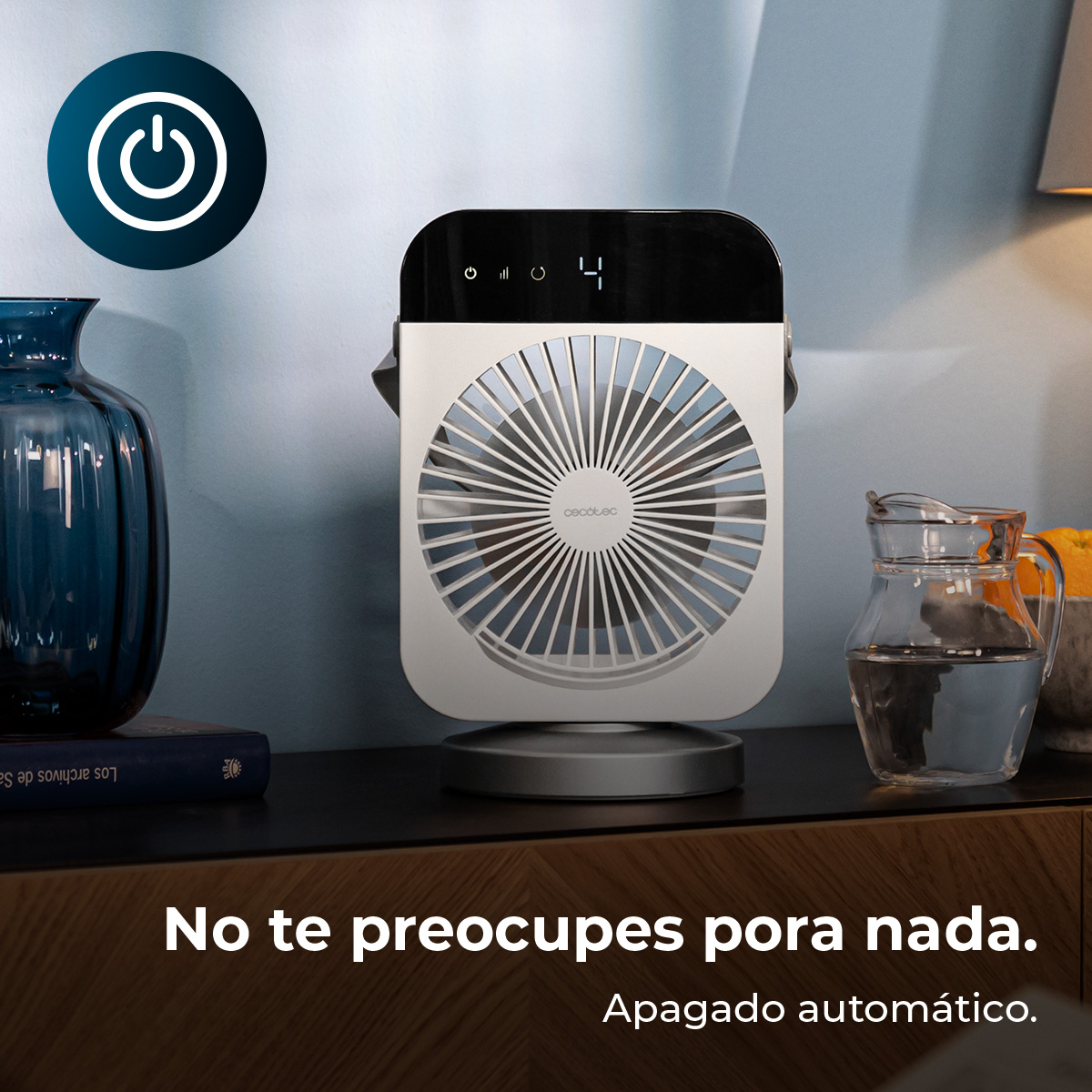 Un ventilador de mesa blanco sobre un mueble de madera junto a un jarrón azul y una jarra de agua.