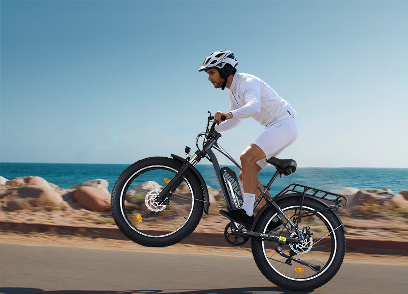 Un ciclista fa un'impennata su un'e-bike lungo una strada costiera con il mare azzurro sullo sfondo.