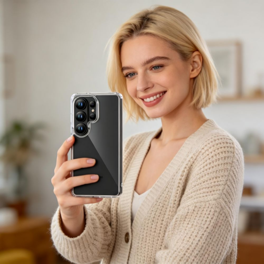 Eine blonde Frau lächelt und hält ein Smartphone in einer klaren Hülle vor sich in einem hellen Raum.