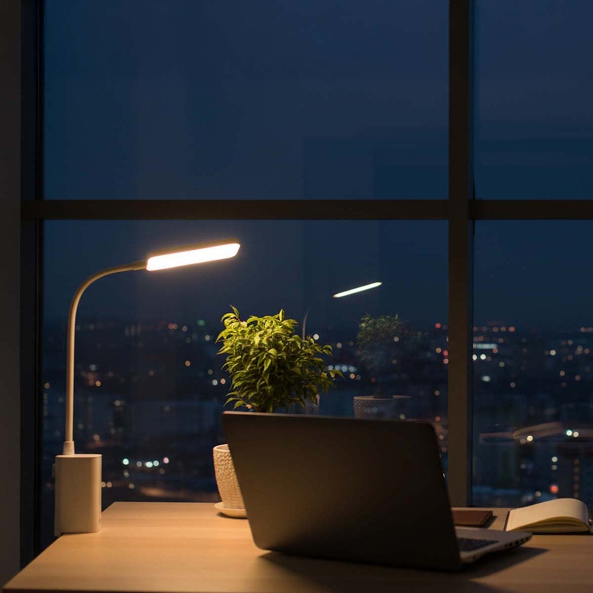 Eine beleuchtete Schreibtischlampe, ein Laptop und Pflanzen vor einem großen Fenster mit nächtlichem Stadtblick.