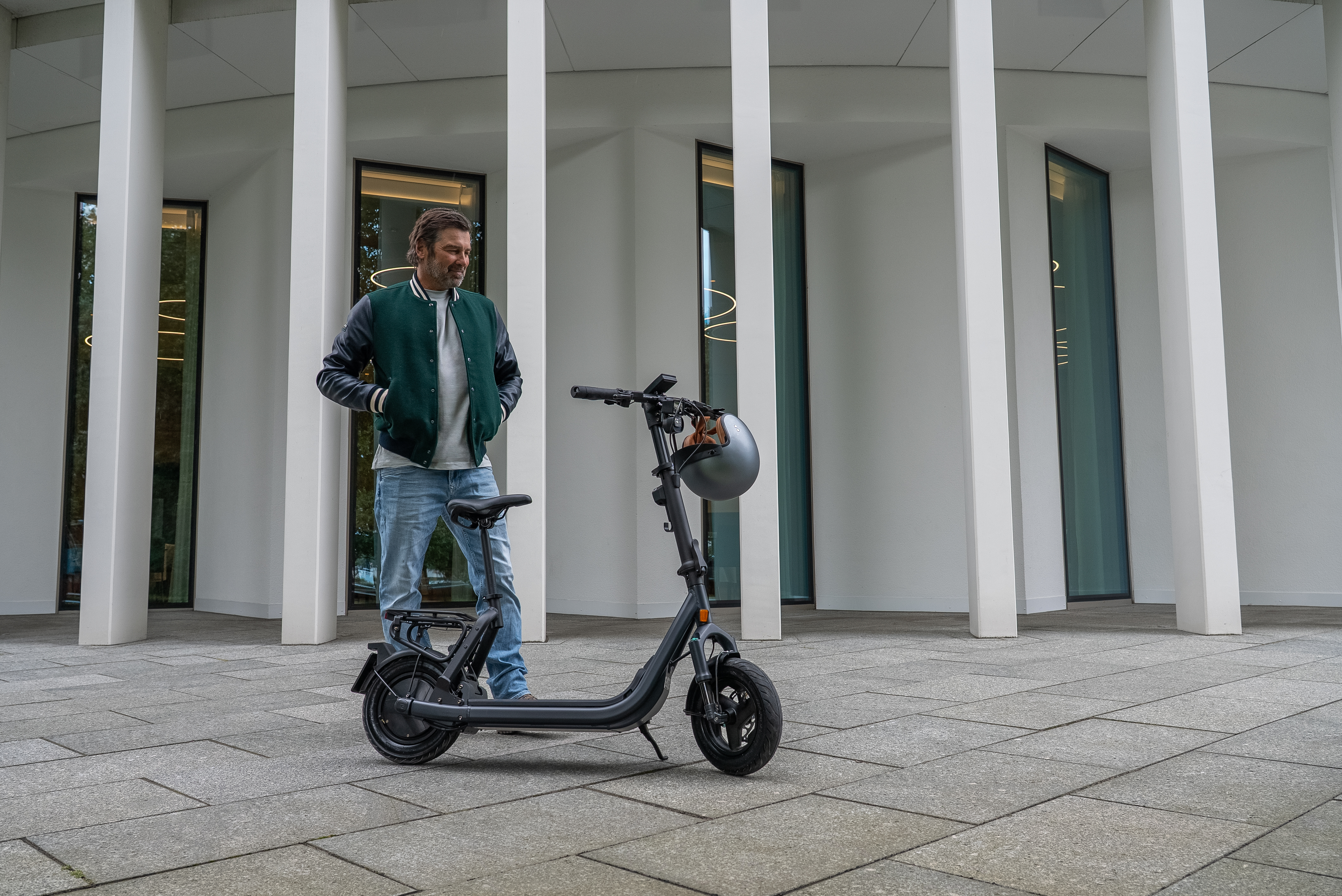 Ein Mann in einer grünen Jacke steht neben einem modernen schwarzen Elektroroller vor einer weißen Säulenarchitektur.