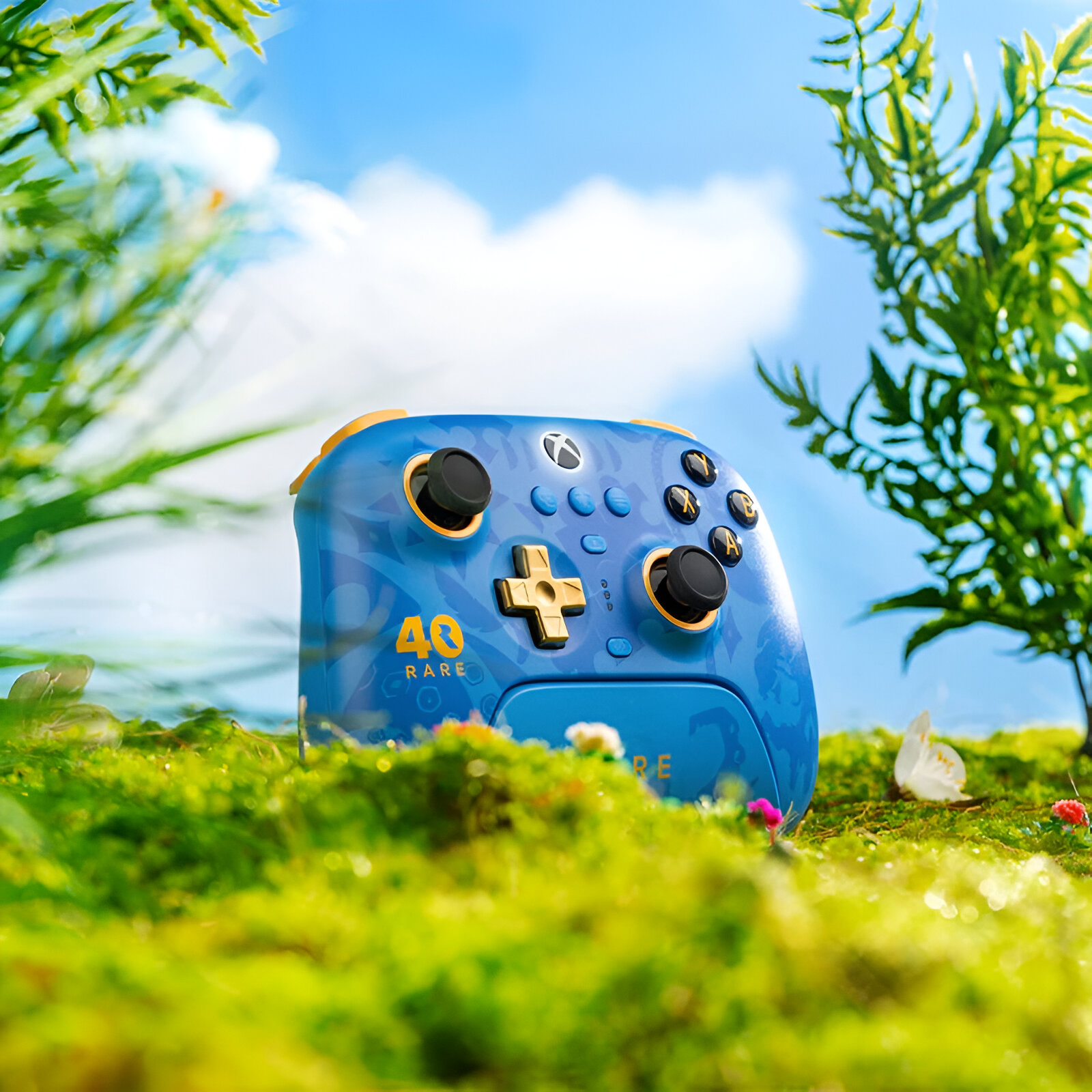 Niebiesko-złoty kontroler Xbox stoi na mchu na tle niebieskiego nieba i zieleni.