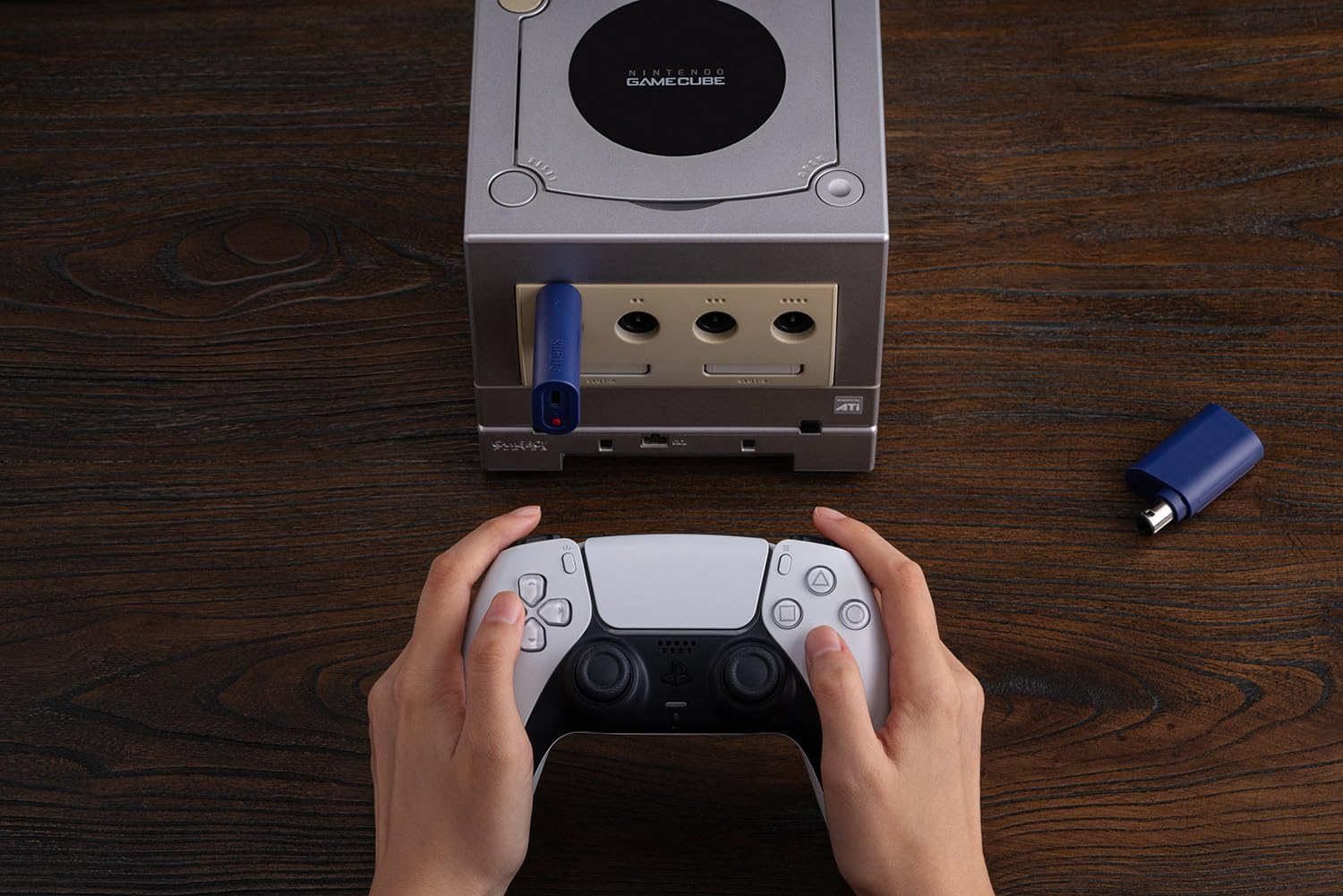 Ręce trzymają biały kontroler PlayStation przed szarym Nintendo GameCube na drewnie.