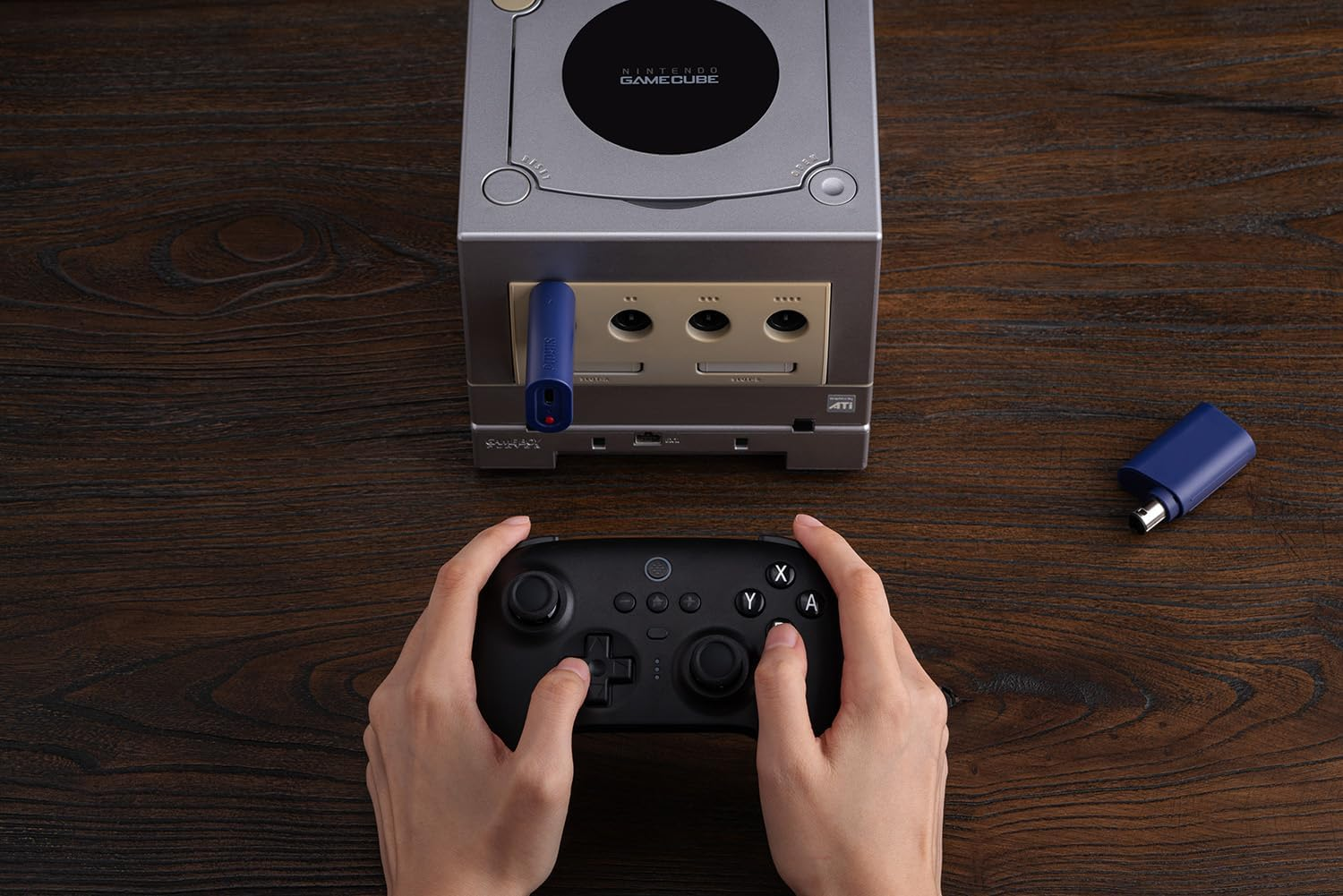 Osoba trzyma czarny kontroler przed srebrną konsolą Nintendo GameCube na drewnianym stole.