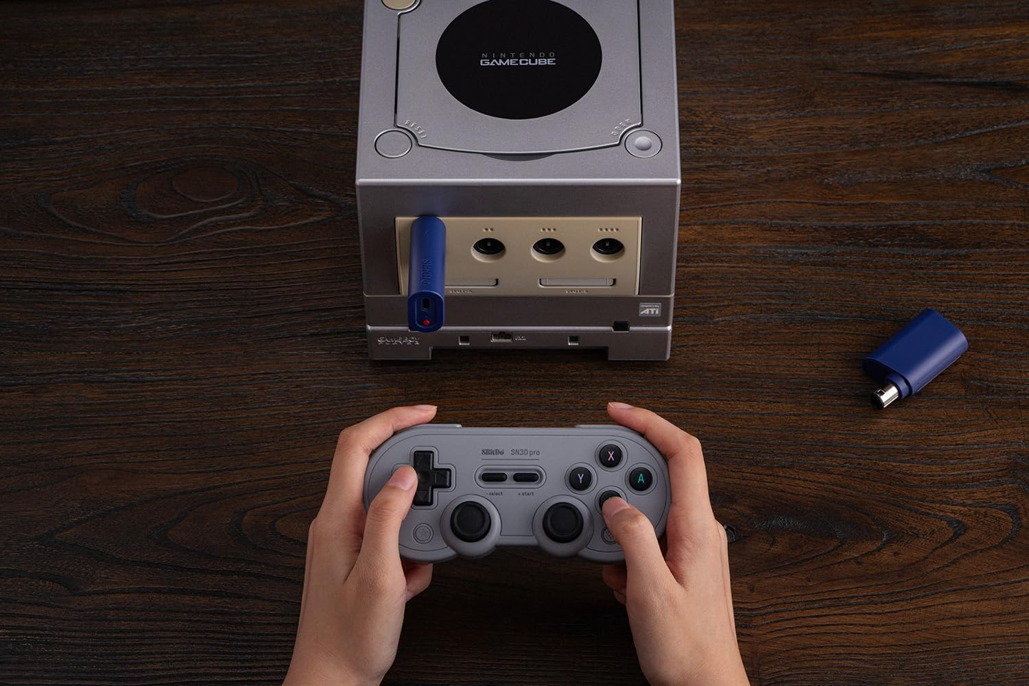Ręce trzymają szary kontroler do gier retro przed konsolą Nintendo GameCube na drewnianym stole.