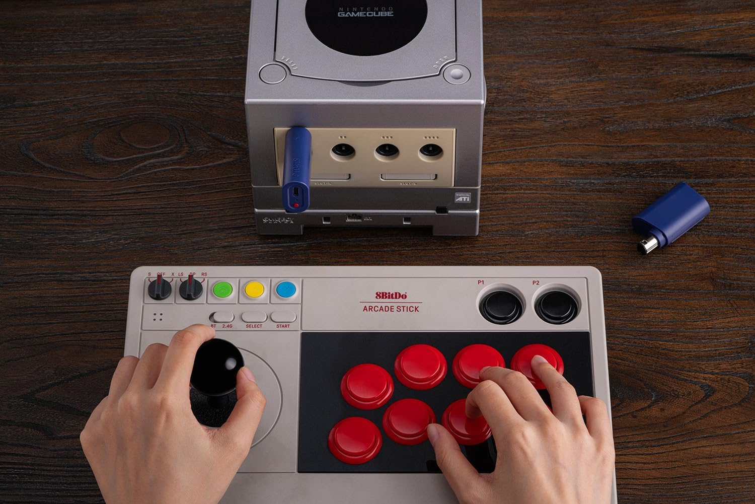 Osoba używa kontrolera arcade 8BitDo na drewnianym stole obok srebrnej konsoli Nintendo GameCube.