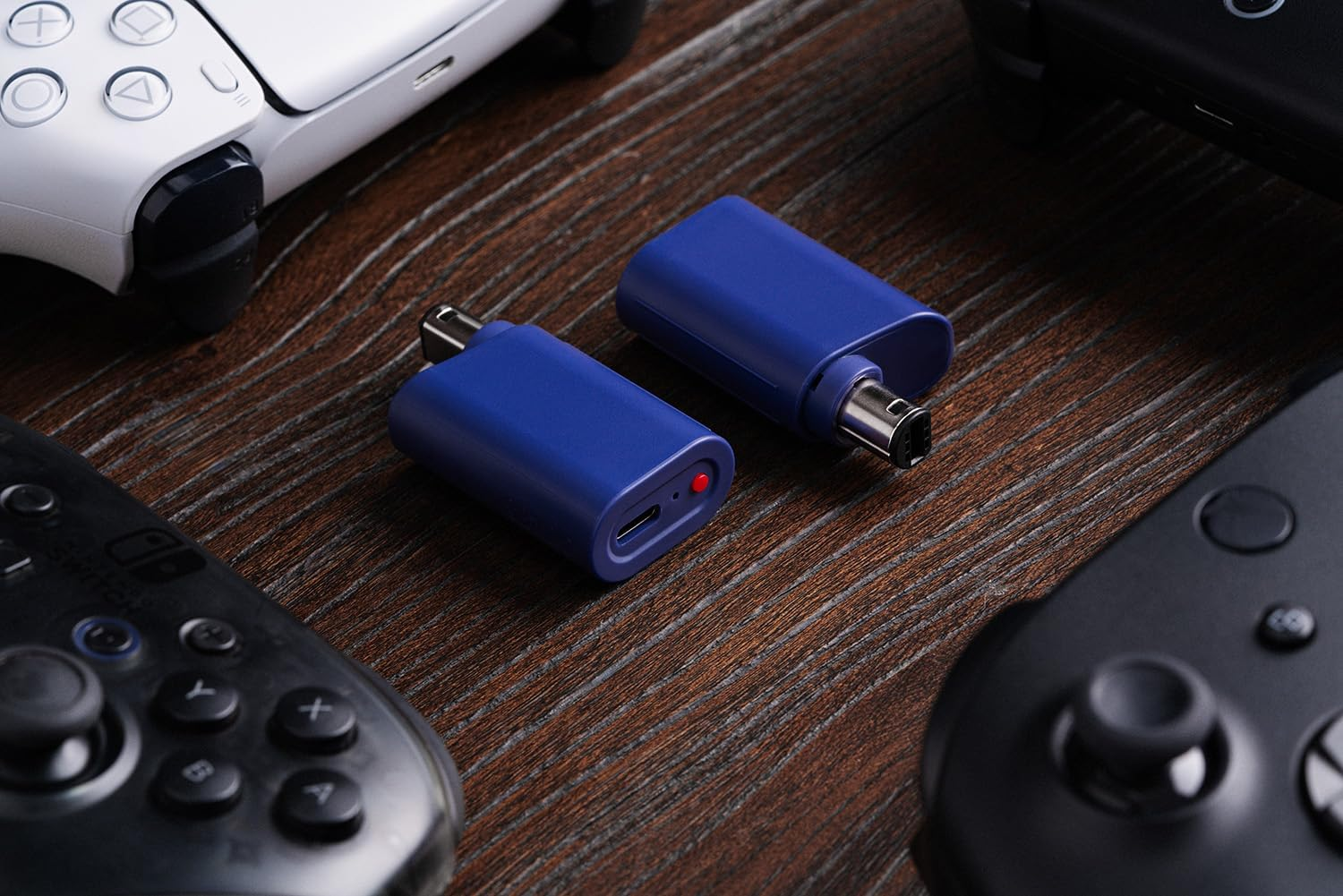 Dwa niebieskie adaptery USB leżą na drewnianej powierzchni w otoczeniu różnych kontrolerów do gier.