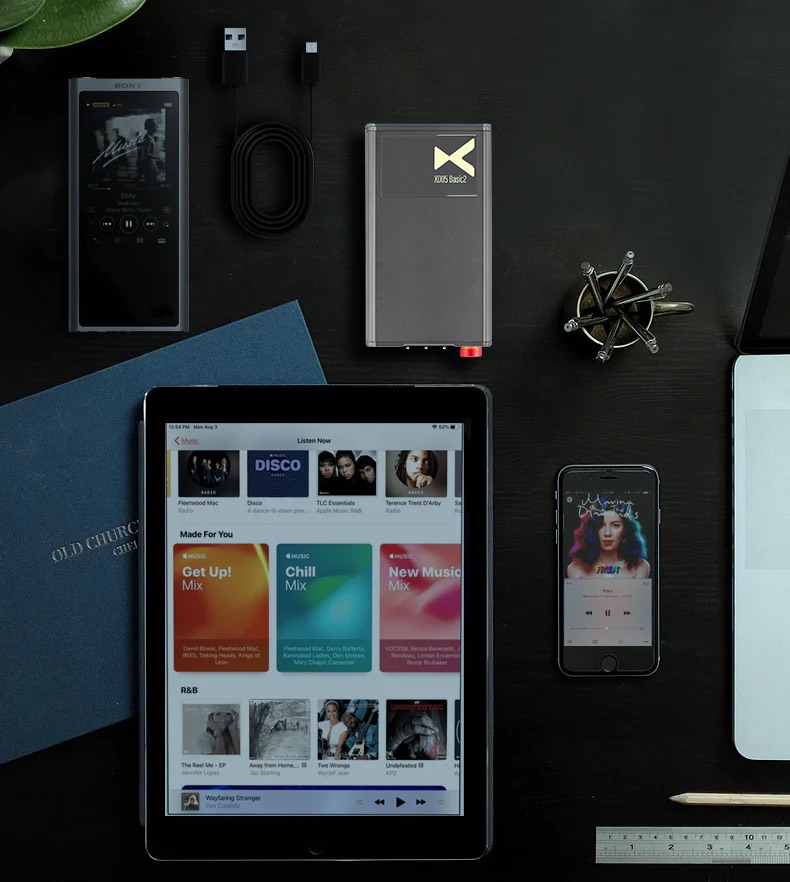 Na ciemnym stole leżą iPad, smartfon, odtwarzacz MP3, słuchawki i notesy.