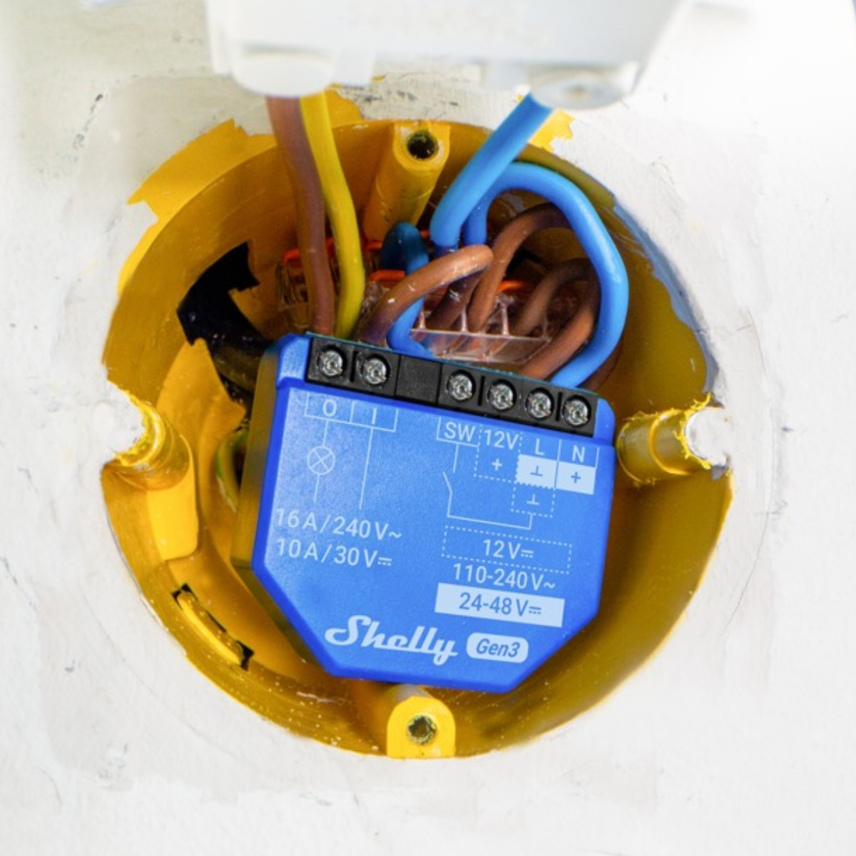 Mavi renkli bir Shelly Gen3 akıllı röle, bağlı elektrik kablolarıyla sarı bir duvar kutusuna monte edilmiştir.