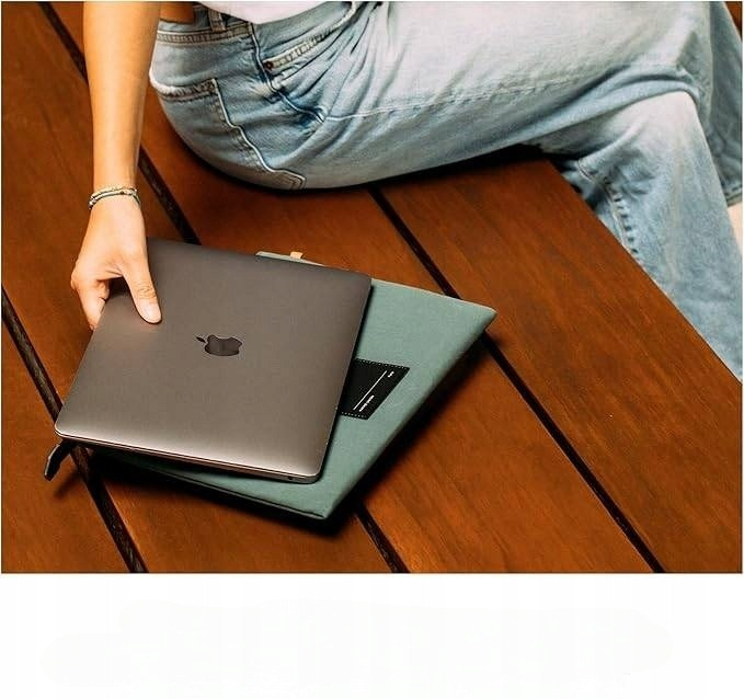 Osoba w dżinsach siedzi na drewnianej ławce, wkładając laptopa do zielonego etui.