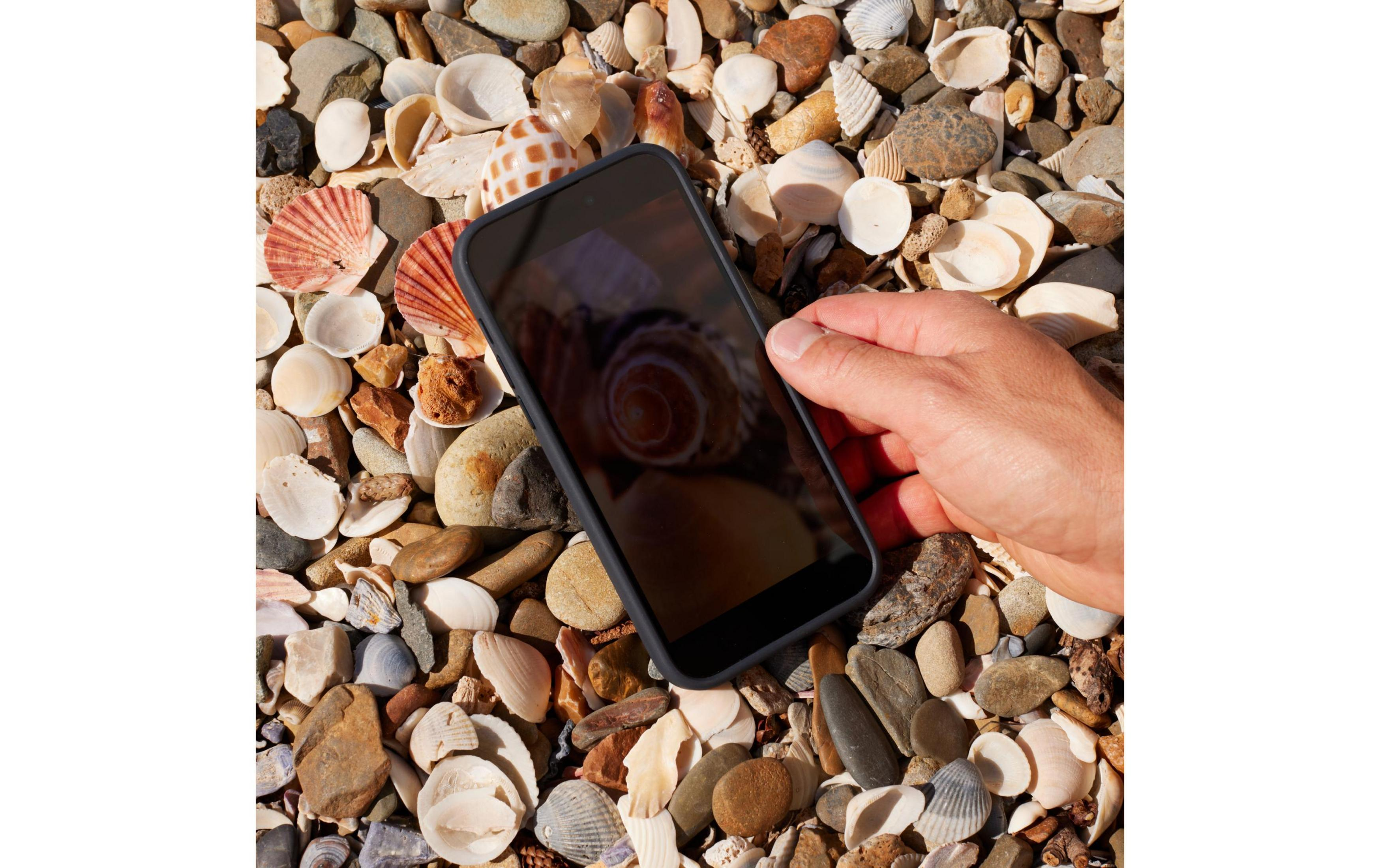 Ręka trzyma smartfon nad plażą pełną muszli i kamyków.