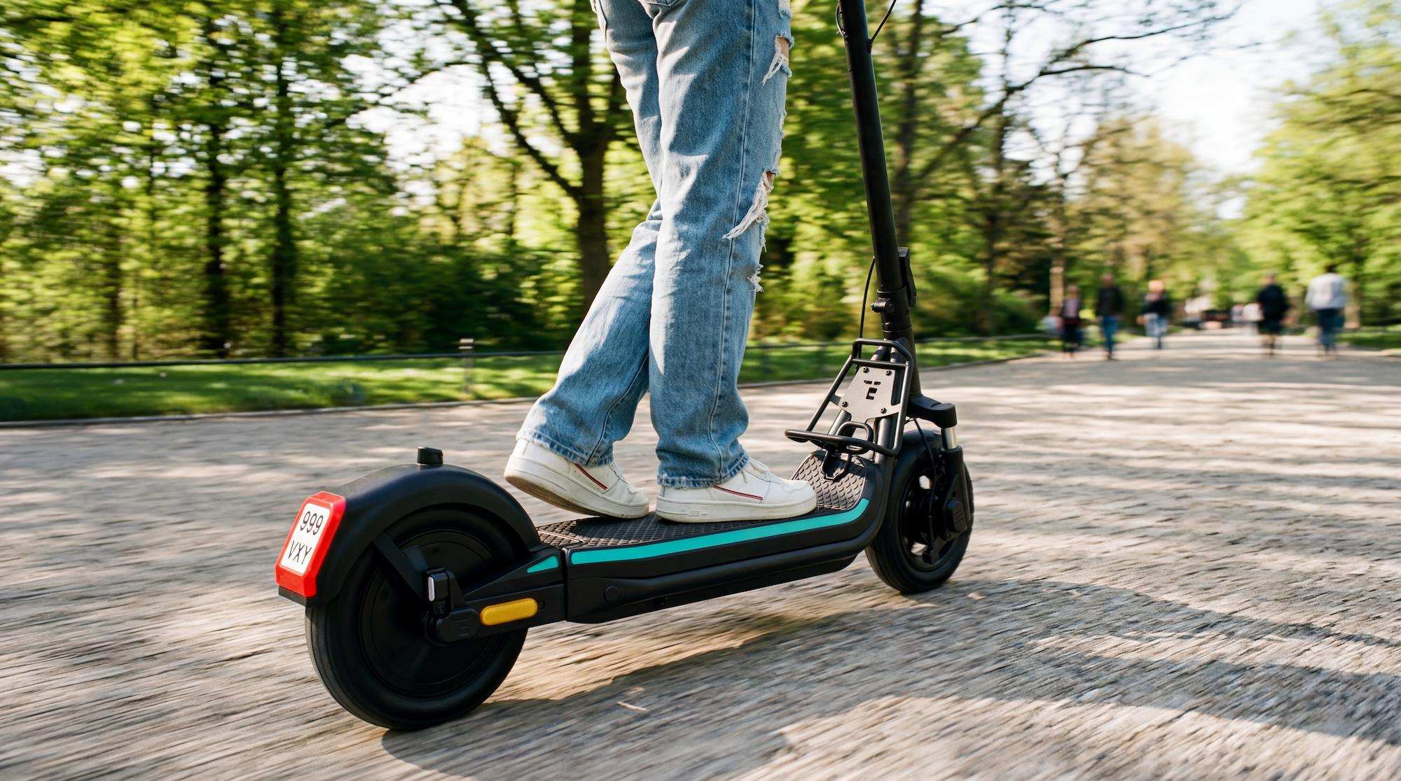 Person in Jeans fährt auf einem schwarzen Elektroroller durch einen Park mit Bäumen im Hintergrund.