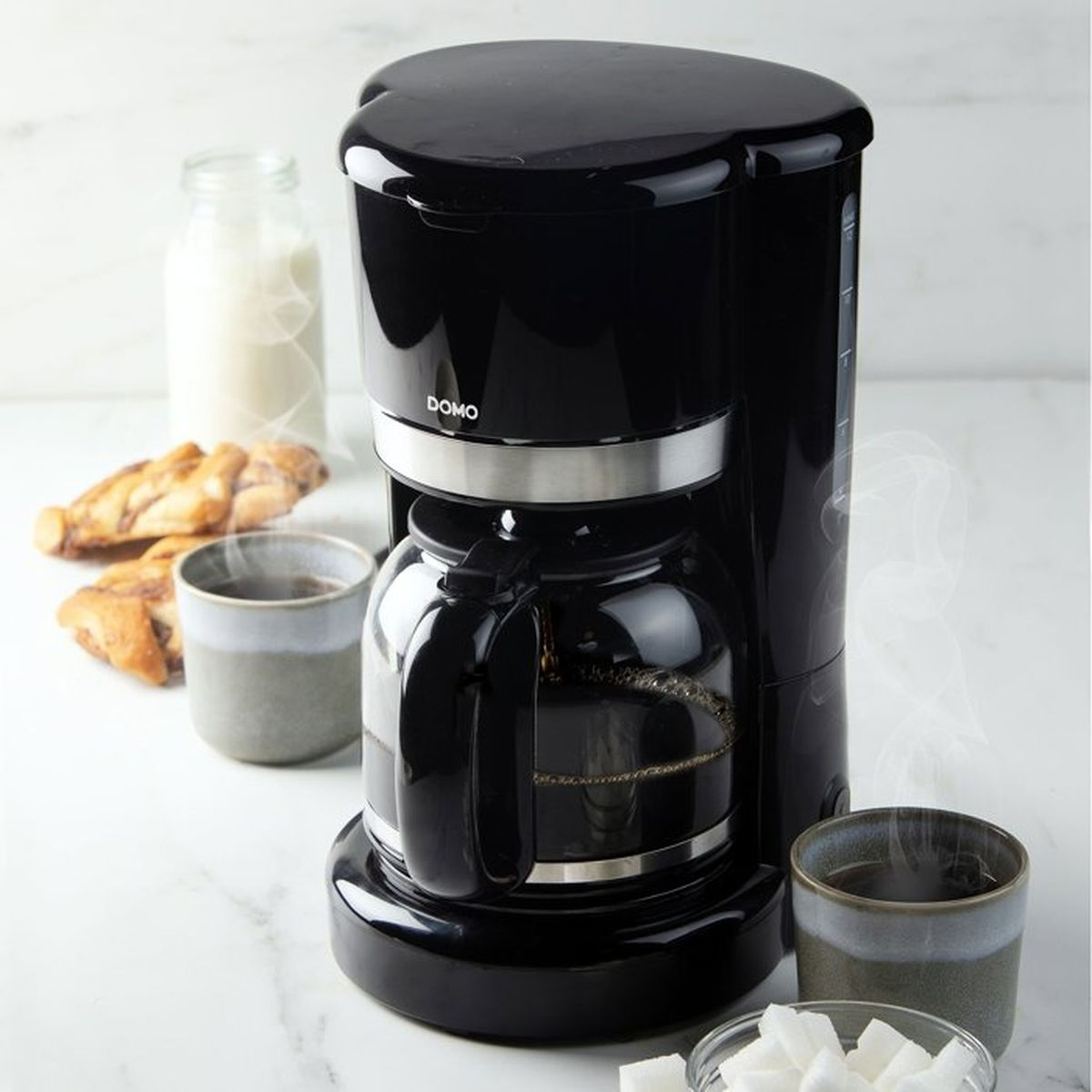 Schwarze Kaffeemaschine auf weißer Marmorplatte mit zwei Tassen, Milch, Zucker und Gebäck.