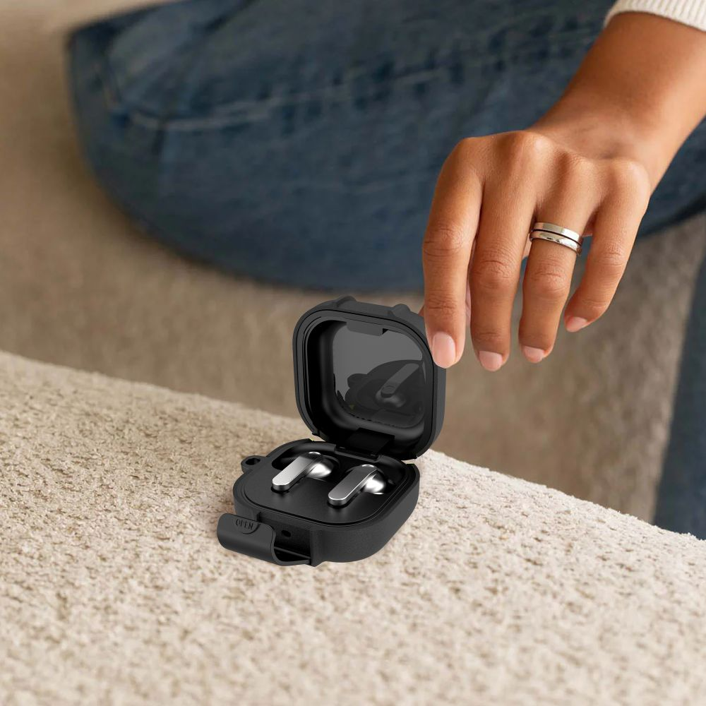 Eine Hand öffnet eine schwarze Ladehülle für silberne Ohrhörer auf einem Sofa.