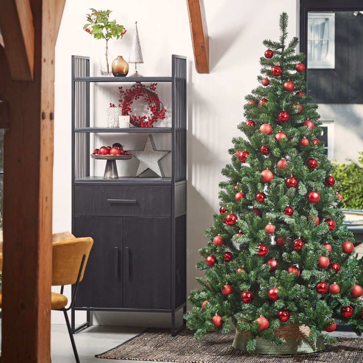 Een kerstboom met rode ballen staat naast een donkere kast met decoratie, in een lichte kamer.