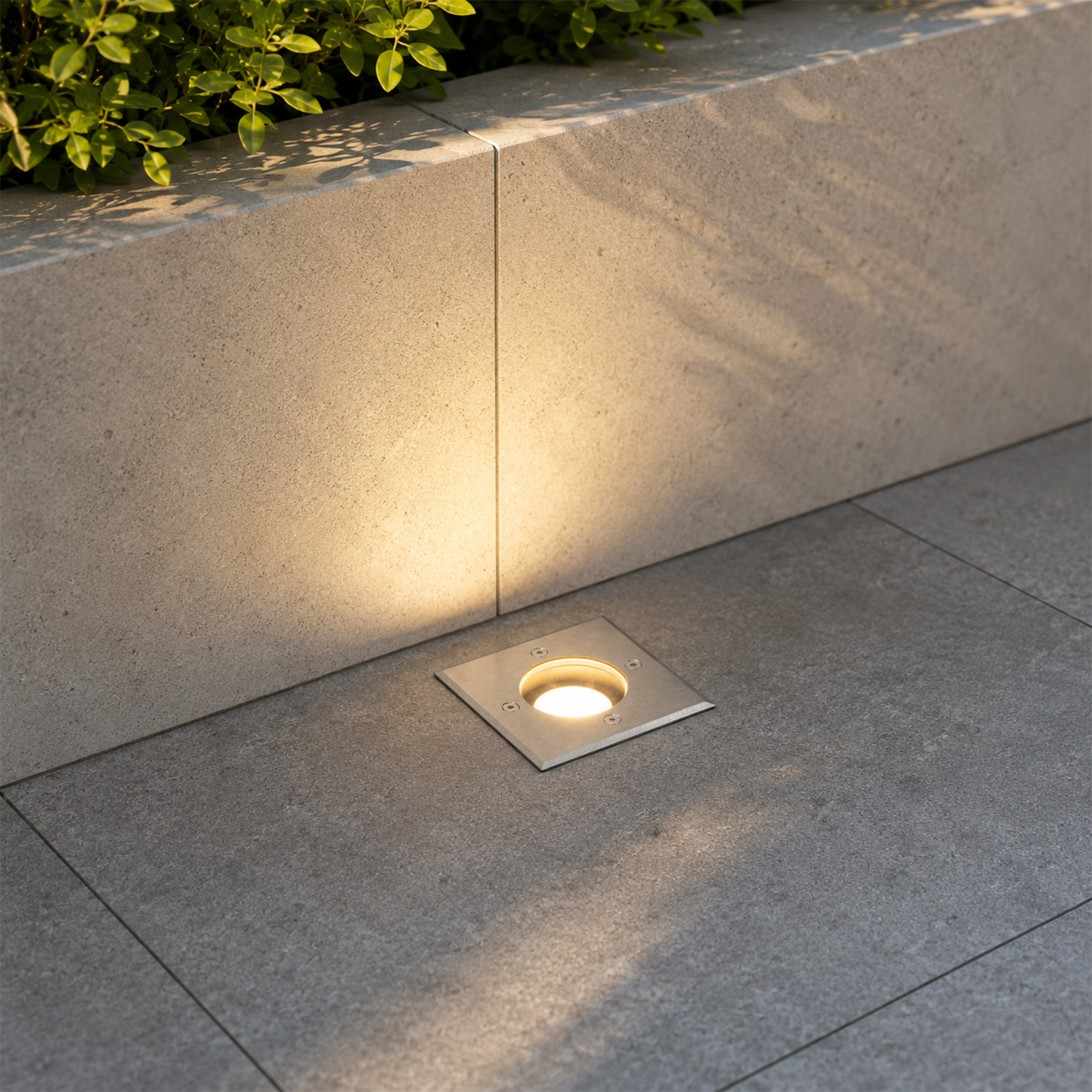 Una luz de suelo cuadrada proyecta una iluminación cálida contra una pared de piedra clara bajo follaje verde.