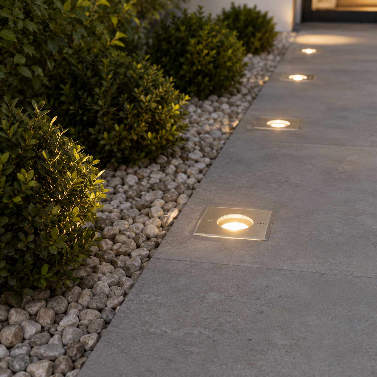 Luces de suelo iluminadas a lo largo de un camino de piedra gris junto a arbustos y guijarros.