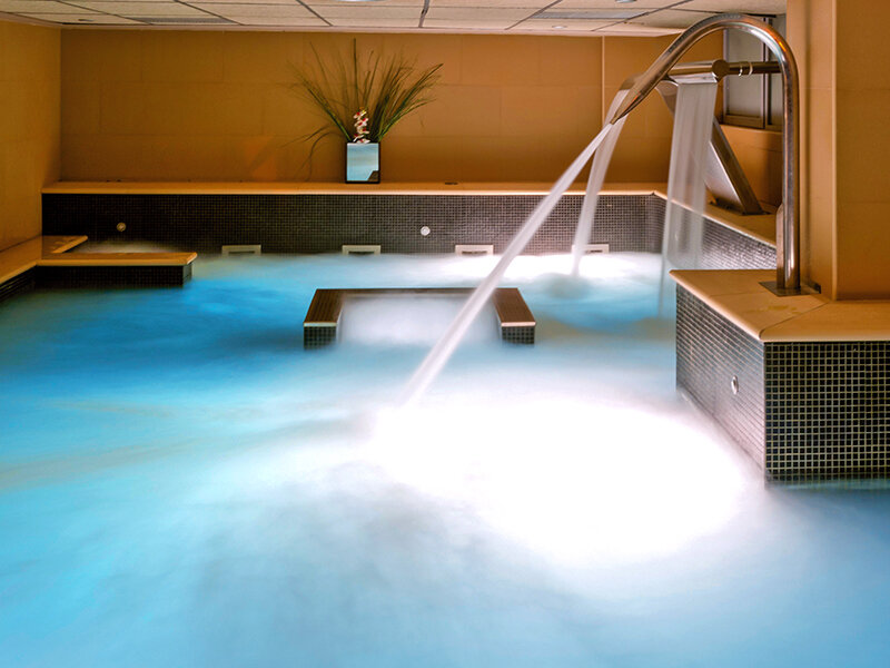 Una piscina de spa interior con agua azul clara, chorros burbujeantes y fuentes de agua metálicas.