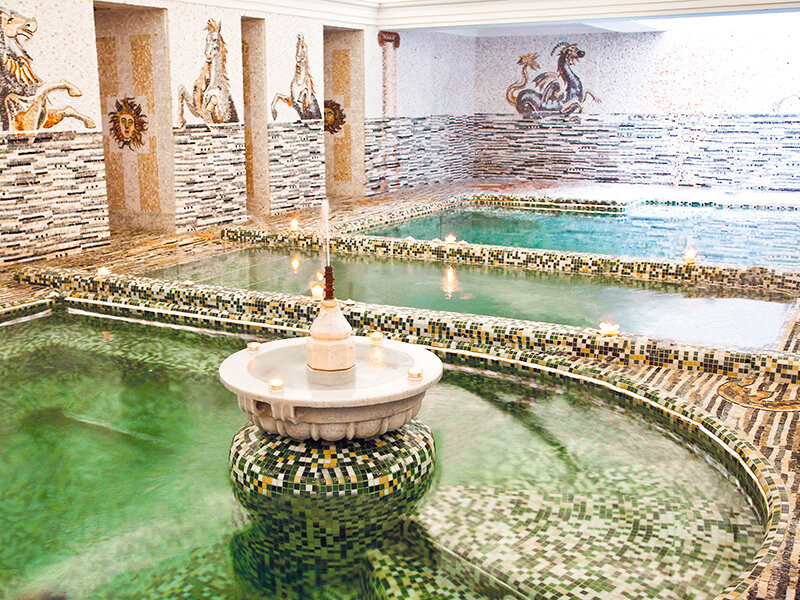 Lujoso baño romano interior con mosaicos, múltiples piscinas y una fuente central.