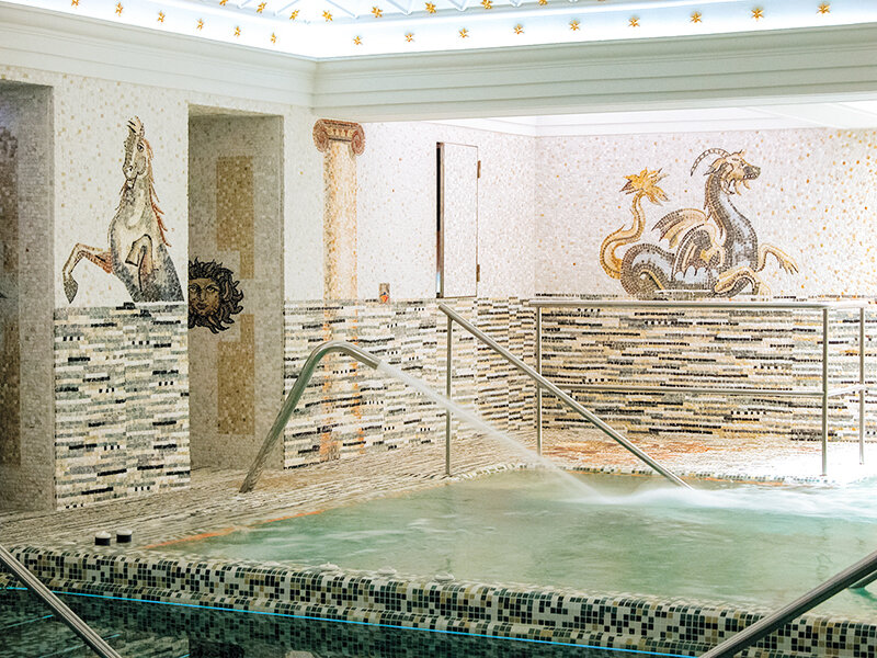 Piscina de lujo con paredes de mosaico, figuras mitológicas y agua clara, equipada con pasamanos de metal.