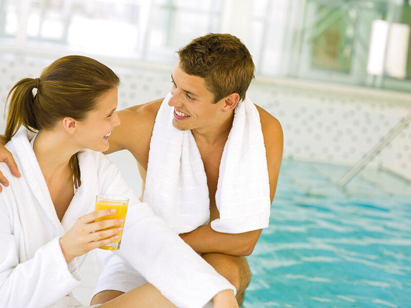 Una pareja en batas blancas junto a la piscina sonríe y bebe zumo de naranja.