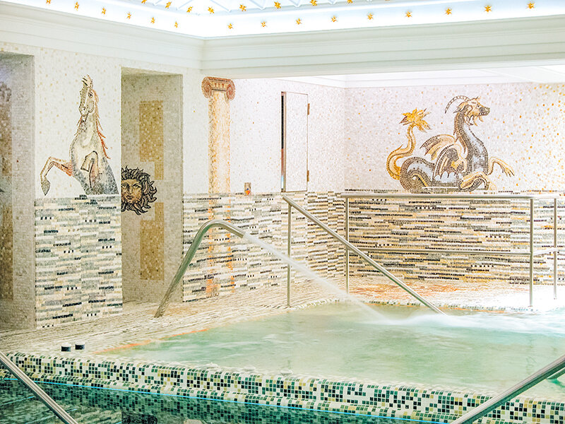 Lujosa piscina romana de interior con paredes de mosaico con un caballo y un dragón, chorros de agua y pasamanos.