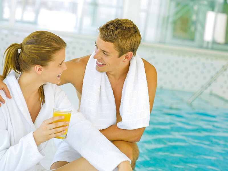 Una pareja sonriente en albornoces blancos junto a una piscina, con zumo de naranja.