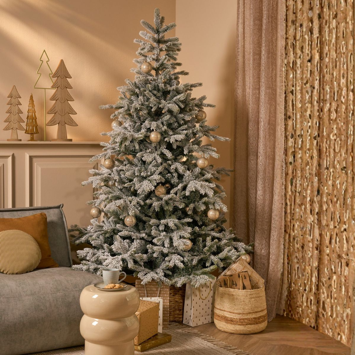 A decorated snow-flocked Christmas tree in a living room next to a gray sofa and gift bags.