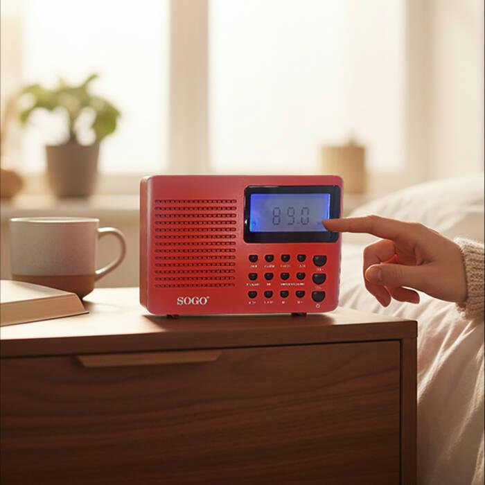 Una mano aziona una radio digitale SOGO rossa su comodino con tazza e pianta sullo sfondo.