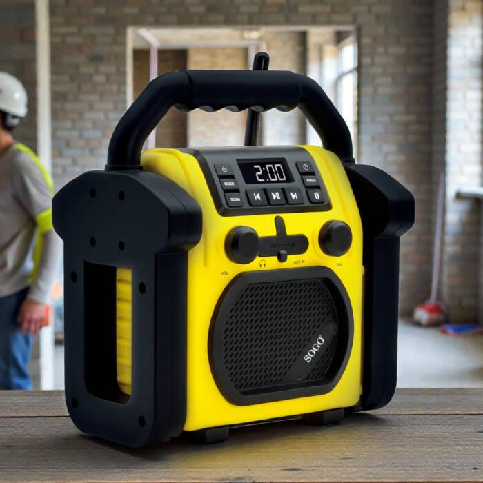 Una radio da cantiere gialla e nera su un tavolo di legno, con un lavoratore nello sfondo sfocato di un cantiere.