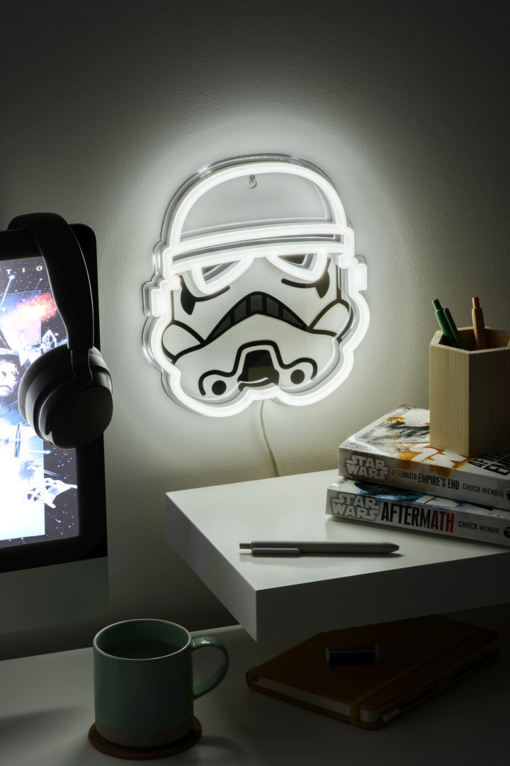 Una luz de neón de Stormtrooper en un escritorio con libros de Star Wars, un monitor, auriculares y una taza verde.