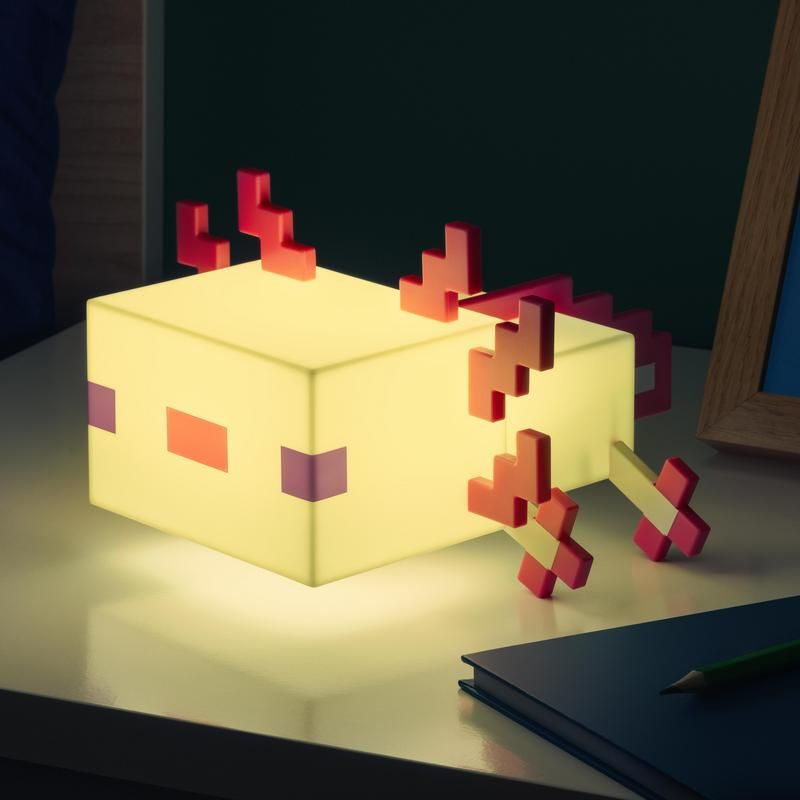 Una lámpara pixelada en forma de ajolote brilla en amarillo con detalles rosados en un escritorio.