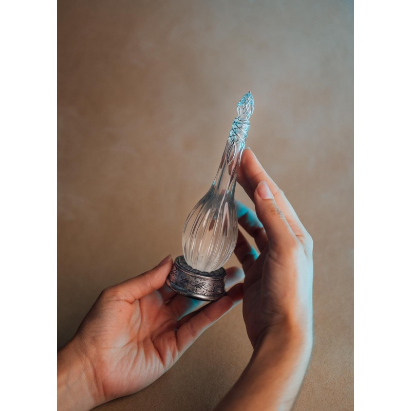 Manos sosteniendo un frasco de perfume de vidrio ornamentado con base metálica sobre fondo marrón.