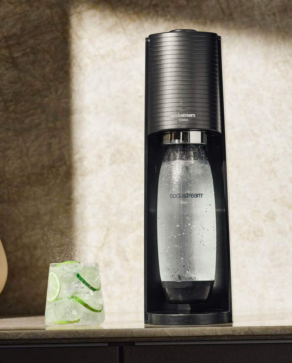 Ein schwarzer Sodastream Terra Wassersprudler steht auf einer Theke neben einem Glas mit Limette und Eis.