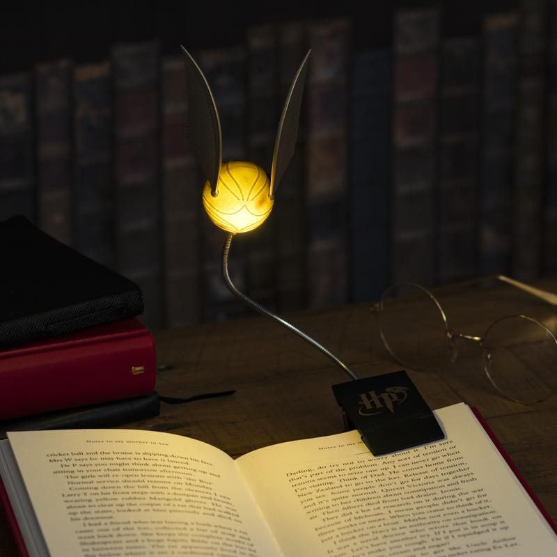 Una luz de lectura de Snitch Dorada unida a un libro abierto, con gafas redondas y libros apilados cerca.