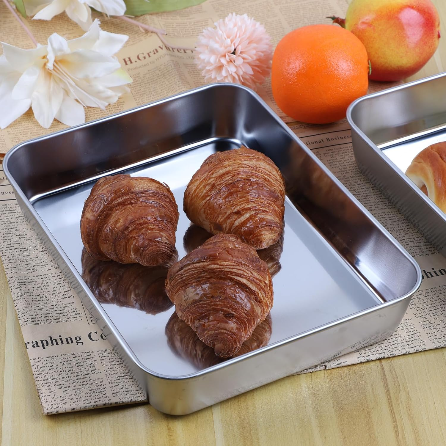 Drei frische Croissants in einem silbernen Metalltablett, umgeben von Obst und Blumen auf Zeitungspapier.