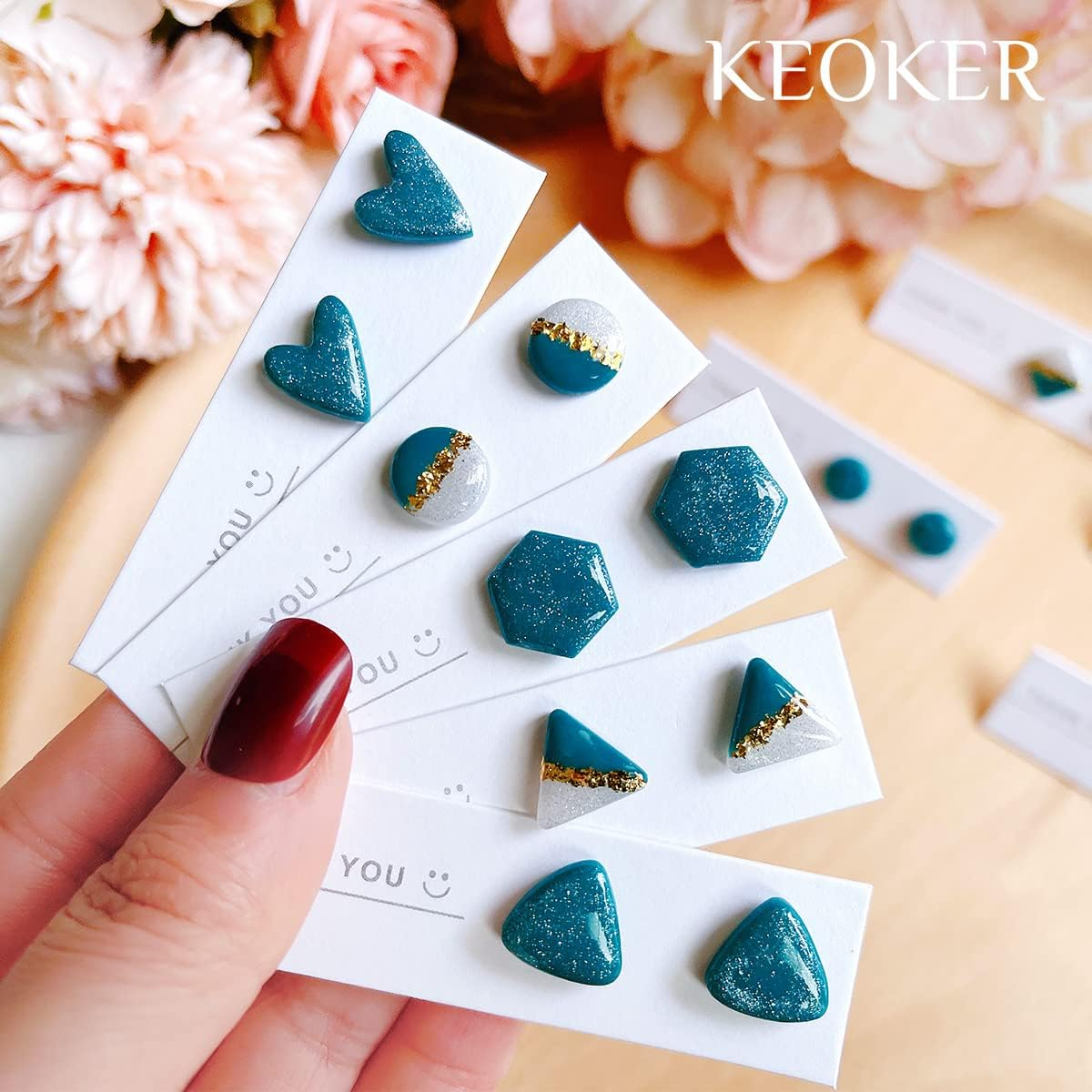 Hand hält Ohrringkarten mit blau-glitzernden ton-Ohrringen in verschiedenen Formen und Goldakzenten vor Blumen.