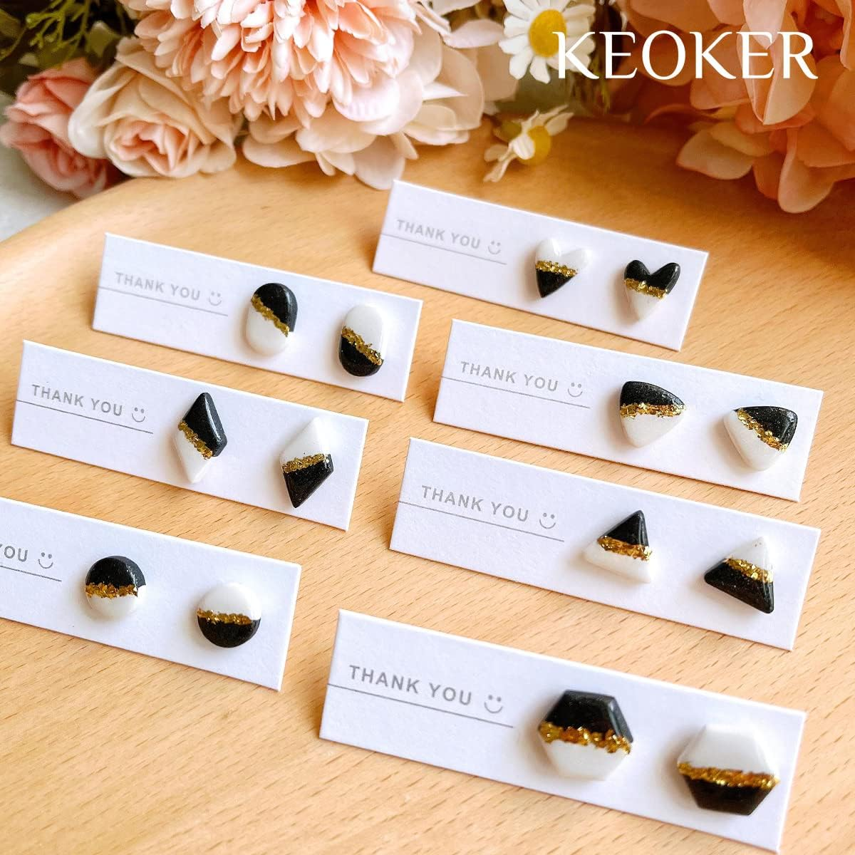 Handgefertigte schwarz-weiße Ohrringe mit Goldfolie auf weißen Thank-You-Karten vor rosa Blumen.