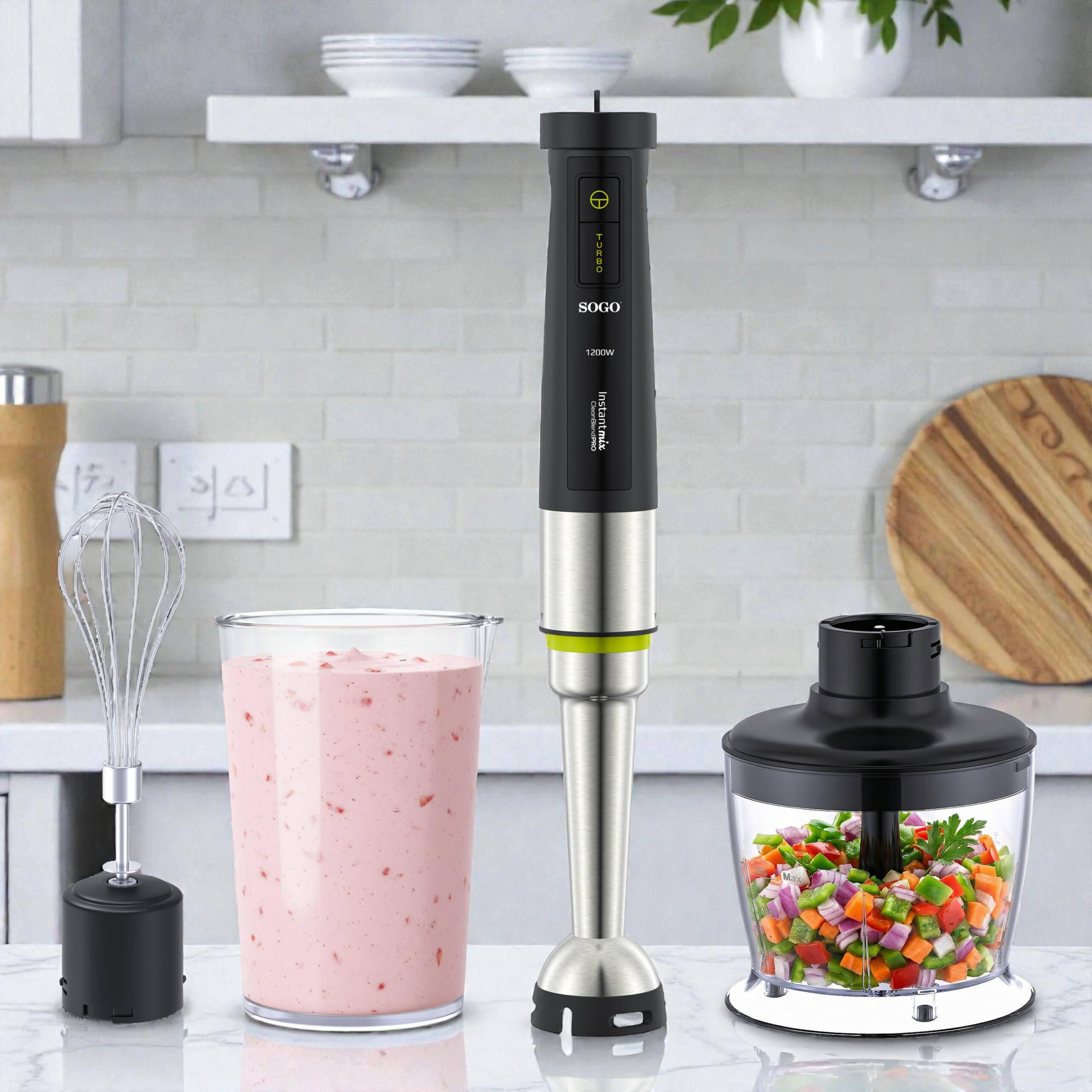Un frullatore ad immersione con frusta, un bicchiere con smoothie e un tritatutto con verdure su un bancone.
